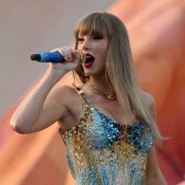 Das Bild zeigt Taylor Swift bei einem Live-Auftritt und dient symbolisch für Artikel über Taylor Swift-Dokus. Sie trägt ein glitzerndes, gold-blaues Bühnenoutfit und singt mit ausdrucksstarkem Gesichtsausdruck in ein blaues Mikrofon. Der verschwommene Hintergrund unterstreicht die emotionale und kraftvolle Atmosphäre der Szene.