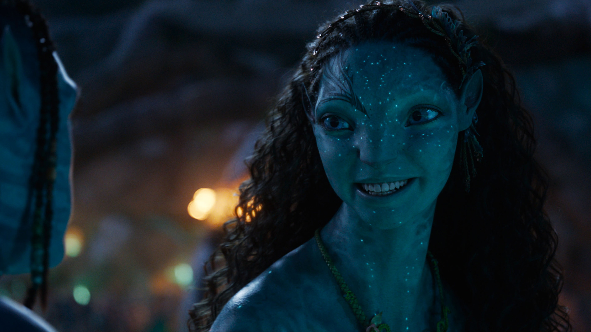 In dieser Szene aus Avatar: Fire and Ash ist die Na'vi-Figur Ronal zu sehen, gespielt von Kate Winslet. Sie lächelt freundlich und wendet sich einer Person gegenüber, die nur teilweise im Bild zu erkennen ist. Ihre Haut leuchtet blau im Licht der Umgebung. Der Alttext dient als Teil einer Avatar: Fire and Ash-Kritik.