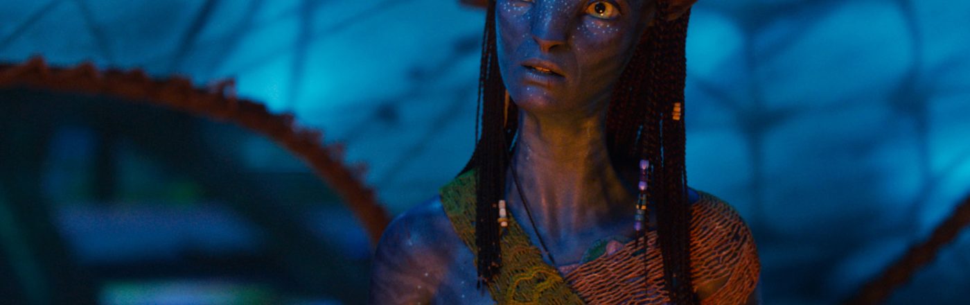 Im Bild aus Avatar: Fire and Ash ist Neytiri, gespielt von Zoe Saldana, in einer nachdenklichen Pose zu sehen. Ihr Gesichtsausdruck ist ernst und wachsam, während sie in einer leuchtend-blauen Umgebung steht. Ihre traditionelle Kleidung und die Zopffrisur unterstreichen ihre kulturelle Identität.