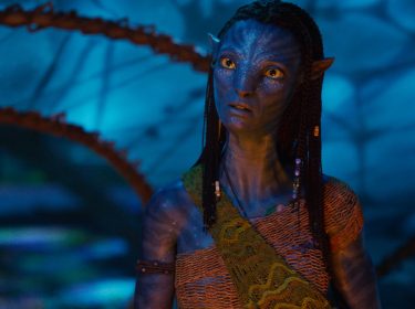 Im Bild aus Avatar: Fire and Ash ist Neytiri, gespielt von Zoe Saldana, in einer nachdenklichen Pose zu sehen. Ihr Gesichtsausdruck ist ernst und wachsam, während sie in einer leuchtend-blauen Umgebung steht. Ihre traditionelle Kleidung und die Zopffrisur unterstreichen ihre kulturelle Identität.