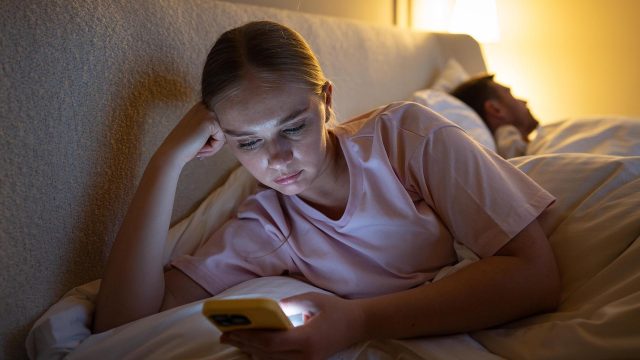 Eine Frau liegt mit einem Mann und Bett und surft mit ihrem Smartphone im Netz.