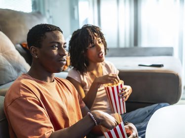 Ein junges Paar sitzt vor dem Sofa auf dem Boden und schaut Fernsehen.