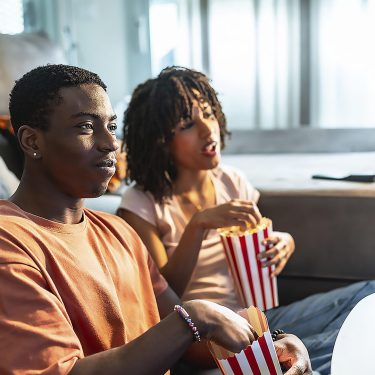Ein junges Paar sitzt vor dem Sofa auf dem Boden und schaut Fernsehen.