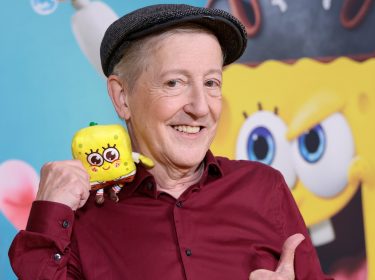 Auf dem Bild ist Santiago Ziesmer, der deutsche SpongeBob-Synchronsprecher, mit einem breiten Lächeln zu sehen. Er trägt ein weinrotes Hemd und eine Schiebermütze und hält ein kleines Stofftier von SpongeBob in der Hand. Im Hintergrund ist eine bunte Wand mit einem großen SpongeBob-Gesicht zu erkennen. Die Stimmung ist fröhlich und verspielt.