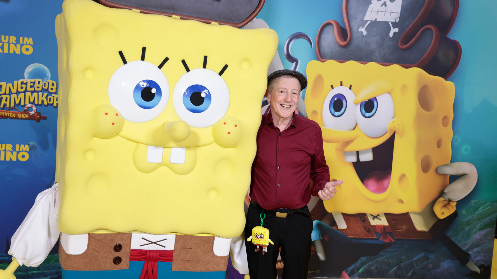Auf dem Bild steht Santiago Ziesmer, der bekannte SpongeBob-Synchronsprecher, lächelnd neben einer lebensgroßen SpongeBob-Figur in Piratenkleidung. Er trägt ein weinrotes Hemd, eine Schiebermütze und hat ein kleines SpongeBob-Plüschtier am Gürtel. Im Hintergrund ist ein farbenfrohes Filmplakat von „SpongeBob Schwammkopf“ zu sehen. Die Stimmung ist heiter und verspielt.