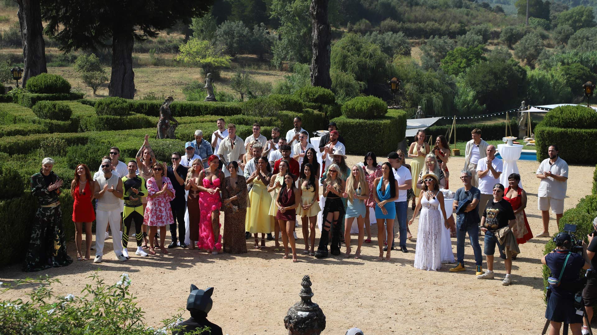 Auf dem Bild zur The 50 Staffel 3 stehen alle 50 Teilnehmer:innen der Reality-Show im Garten einer mediterranen Villa. Die Gruppe ist bunt gemischt – mit auffälligen Outfits, sommerlicher Kleidung und fröhlicher Stimmung. Im Vordergrund ist die ikonische Spielfigur mit Katzenmaske zu sehen. Die Kandidat:innen klatschen und wirken gespannt auf das bevorstehende Spiel.