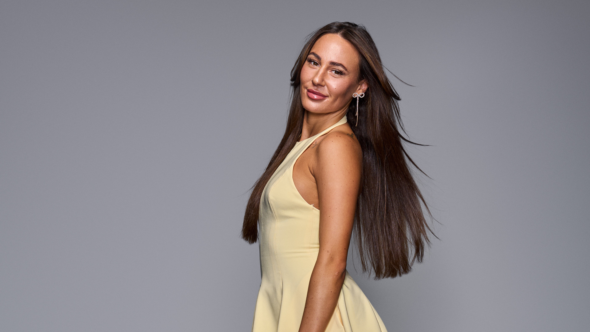 Auf dem Bild zur The 50 Staffel 3 ist Alessia Herren zu sehen. Sie trägt ein hellgelbes, ärmelloses Kleid und schaut mit einem leichten Lächeln über die Schulter in die Kamera. Ihr glattes, braunes Haar fällt locker über den Rücken, während dezenter Schmuck an ihrem Ohr glänzt. Der Hintergrund ist schlicht grau gehalten, was den Fokus auf ihre elegante Erscheinung lenkt.