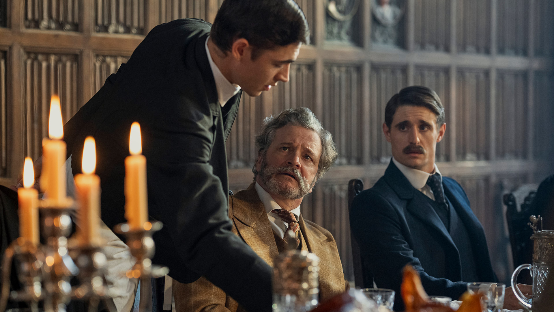 Auf diesem Bild aus Young Sherlock ist Sherlock Holmes, gespielt von Hero Fiennes Tiffin, links im Bild zu sehen, während er sich zu einem älteren Herrn vorbeugt. In der Mitte sitzt Colin Firth in einer beigefarbenen Anzugjacke und blickt ihn ernst an. Im Hintergrund sitzt Dónal Finn mit zurückgekämmten Haaren und Schnurrbart, in dunklem Anzug. Die Stimmung ist angespannt, das Setting wirkt historisch und formell mit dunkler Holzvertäfelung und Kerzenschein.