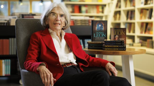 Donna Leon sitzt in einem Sessel und lächelt freundlich in die Kamera. Daneben steht ein kleiner Tisch mit einigen ihrer Bücher darauf.