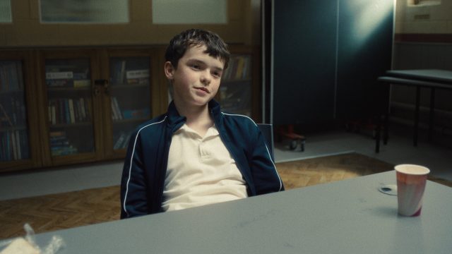 In den besten Serien 2025 siehst du eine Szene aus Adolescence. Owen Cooper als Jamie Miller sitzt in einem Raum mit Bücherregal im Hintergrund an einem grauen Tisch. Er trägt ein weißes Poloshirt und eine dunkelblaue Trainingsjacke. Auf dem Tisch steht ein Pappbecher.