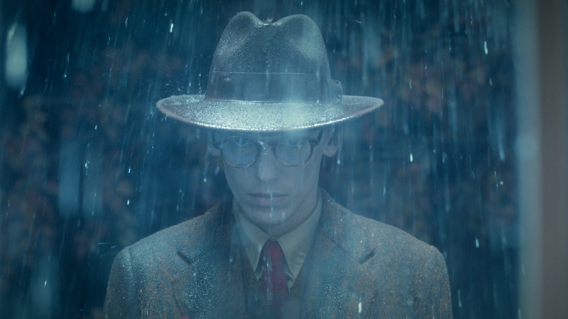 Bei Stranger Things Staffel 5 siehst du Henry Creel, gespielt von Jamie Campbell Bower, in einer düsteren Regenszene. Er trägt einen nassen Fedora-Hut und eine altmodische Brille, sein Blick ist ernst und geheimnisvoll.
