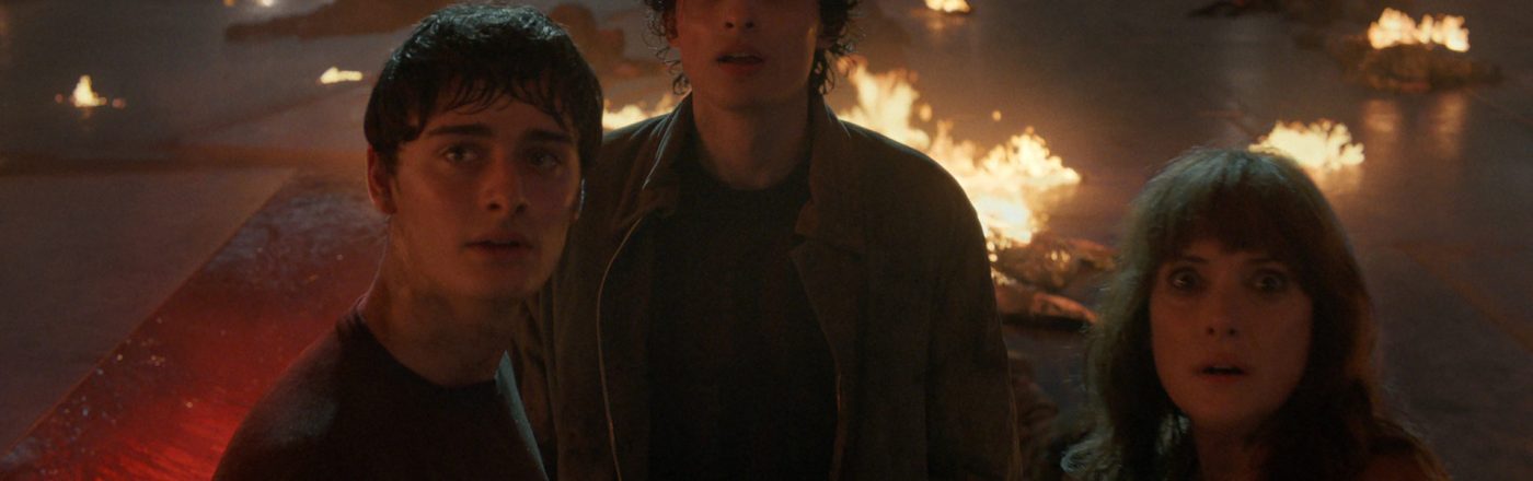 Bei Stranger Things Staffel 5 siehst du Will Byers (Noah Schnapp), Mike Wheeler (Finn Wolfhard) und Joyce Byers (Winona Ryder) inmitten einer dramatischen Szene voller Flammen. Die drei Charaktere blicken erschrocken nach oben, umgeben von brennenden Trümmern