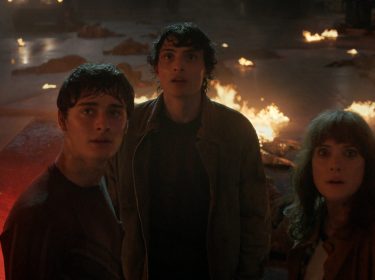 Bei Stranger Things Staffel 5 siehst du Will Byers (Noah Schnapp), Mike Wheeler (Finn Wolfhard) und Joyce Byers (Winona Ryder) inmitten einer dramatischen Szene voller Flammen. Die drei Charaktere blicken erschrocken nach oben, umgeben von brennenden Trümmern