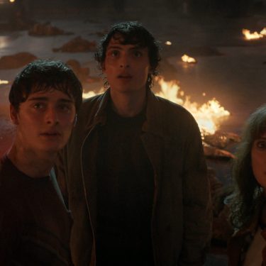 Bei Stranger Things Staffel 5 siehst du Will Byers (Noah Schnapp), Mike Wheeler (Finn Wolfhard) und Joyce Byers (Winona Ryder) inmitten einer dramatischen Szene voller Flammen. Die drei Charaktere blicken erschrocken nach oben, umgeben von brennenden Trümmern