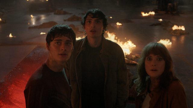 Bei Stranger Things Staffel 5 siehst du Will Byers (Noah Schnapp), Mike Wheeler (Finn Wolfhard) und Joyce Byers (Winona Ryder) inmitten einer dramatischen Szene voller Flammen. Die drei Charaktere blicken erschrocken nach oben, umgeben von brennenden Trümmern