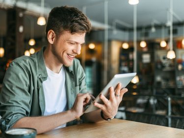 Ein junger Mann sitzt in einer Buchhandlung und schaut auf ein Tablet.