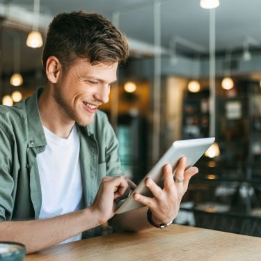 Ein junger Mann sitzt in einer Buchhandlung und schaut auf ein Tablet.