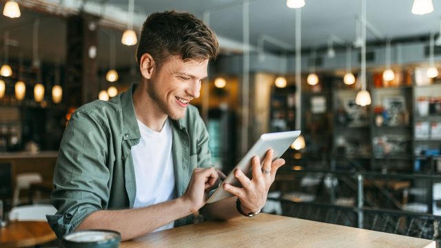 Ein junger Mann sitzt in einer Buchhandlung und schaut auf ein Tablet.
