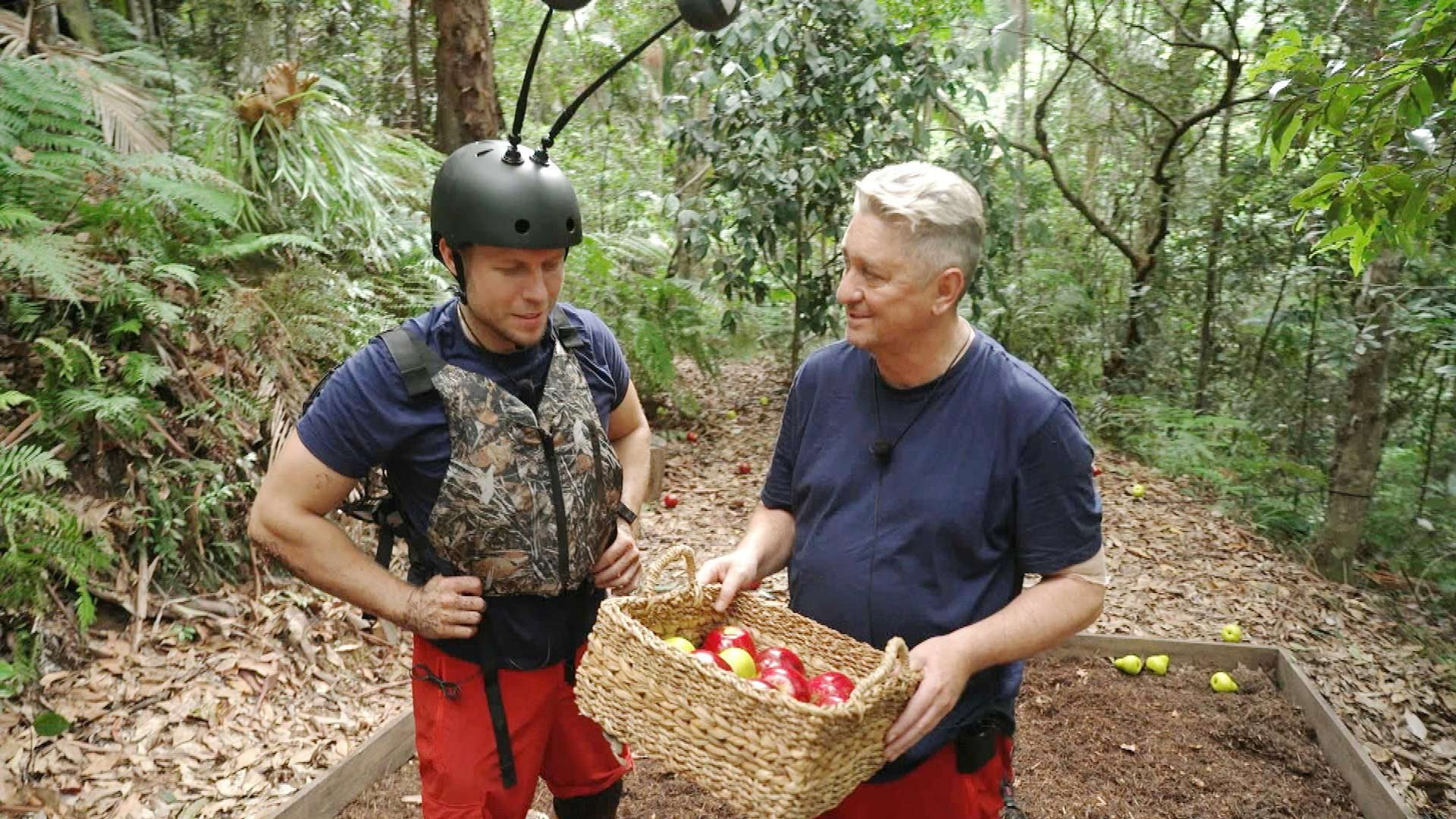 Auf dem Bild zur Dschungelcamp 2026 Schatzsuche „Die Dschungel-Ernte“ sind Patrick und Hubert im australischen Dschungel zu sehen. Patrick trägt einen Schutzhelm mit Antennen und eine Weste, während Hubert einen Korb mit roten und grünen Äpfeln hält. Beide wirken konzentriert und ruhig. Die Stimmung ist kooperativ, angespannt und von Teamarbeit geprägt.