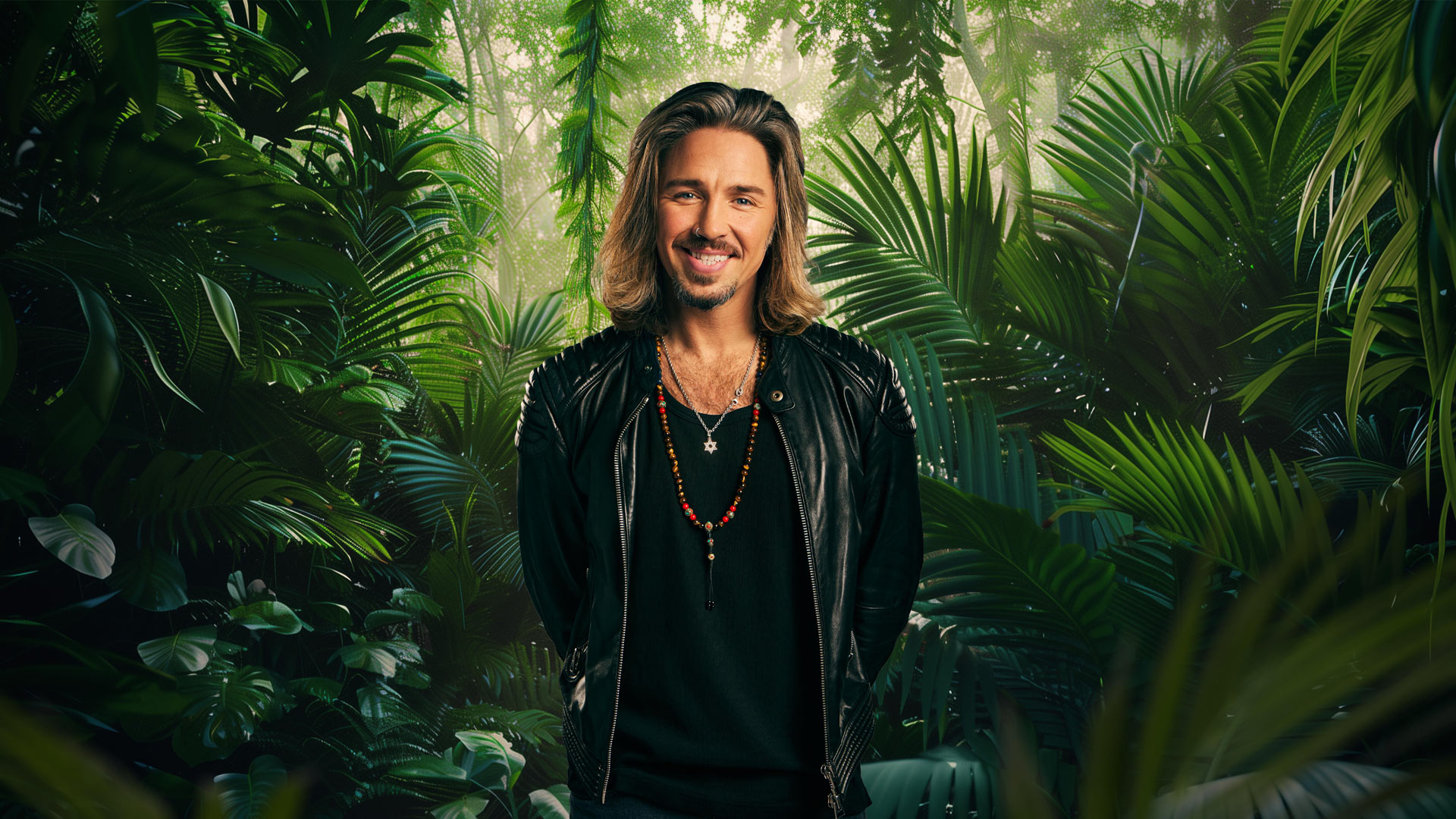 Auf dem Bild zum Dschungelcamp 2026 steht Teilnehmer Gil Ofarim vor einem dichten, grünen Dschungelhintergrund. Er trägt eine schwarze Lederjacke über einem dunklen Shirt und mehrere auffällige Ketten, darunter eine mit Davidstern. Mit schulterlangem Haar, lässigem Lächeln und charismatischer Ausstrahlung wirkt er entspannt, aber kampfbereit für das Abenteuer.