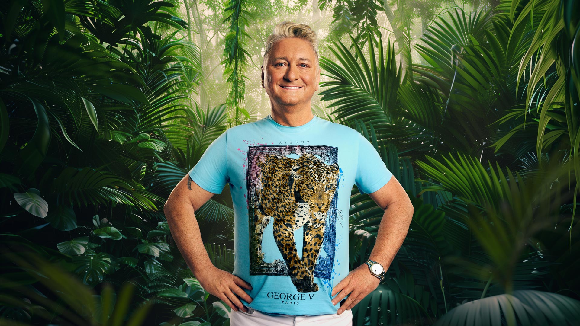 Auf dem Bild zum Dschungelcamp 2026 steht Teilnehmer Hubert Fella vor dichtem, tropischem Dschungelgrün. Er trägt ein auffälliges, hellblaues T-Shirt mit einem großen Gepardenmotiv der Marke George V Paris und kombiniert es mit einer weißen Hose. Mit beiden Händen in der Hüfte und einem breiten Lächeln zeigt er sich farbenfroh, offen und gut gelaunt.