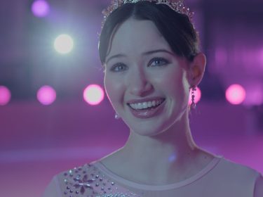 Madelyn Keys in Finding Her Edge: Eine junge Eiskunstläuferin mit Diadem lächelt im Schein bunter Lichter in der Eishalle.