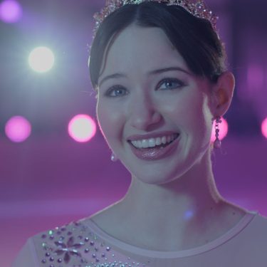 Madelyn Keys in Finding Her Edge: Eine junge Eiskunstläuferin mit Diadem lächelt im Schein bunter Lichter in der Eishalle.