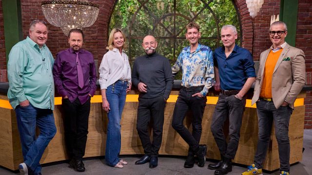 Auf dem Bild sind die Bares für Rares Händler Walter Lehnertz, Wolfgang Pauritsch, Susanne Steiger, Fabian Kahl, Christian Velten und Anaisio Guedes gemeinsam mit Moderator Horst Lichter zu sehen. Sie stehen gut gelaunt im Studio der ZDF-Sendung, vor einer Kulisse mit rustikaler Backsteinwand und großen Fenstern mit Blick ins Grüne.