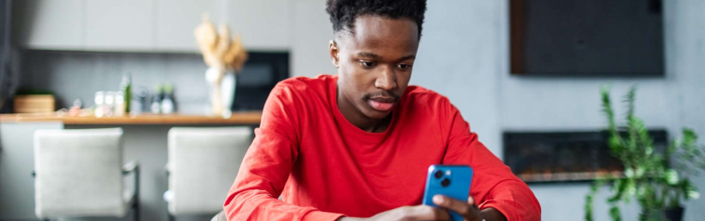 Ein junger Mann sitzt in einer Wohnküche an einem Tisch und schaut auf sein Smartphone.