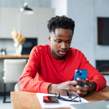 Ein junger Mann sitzt in einer Wohnküche an einem Tisch und schaut auf sein Smartphone.