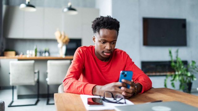 Ein junger Mann sitzt in einer Wohnküche an einem Tisch und schaut auf sein Smartphone.