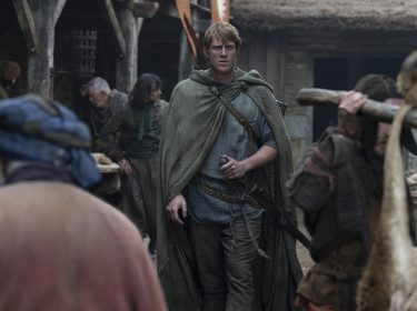 In dieser Szene aus A Knight of the Seven Kingdoms ist ein junger Mann mit blondem Haar und Umhang in einem Dorf zu sehen.