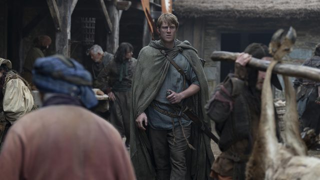 In dieser Szene aus A Knight of the Seven Kingdoms ist ein junger Mann mit blondem Haar und Umhang in einem Dorf zu sehen.