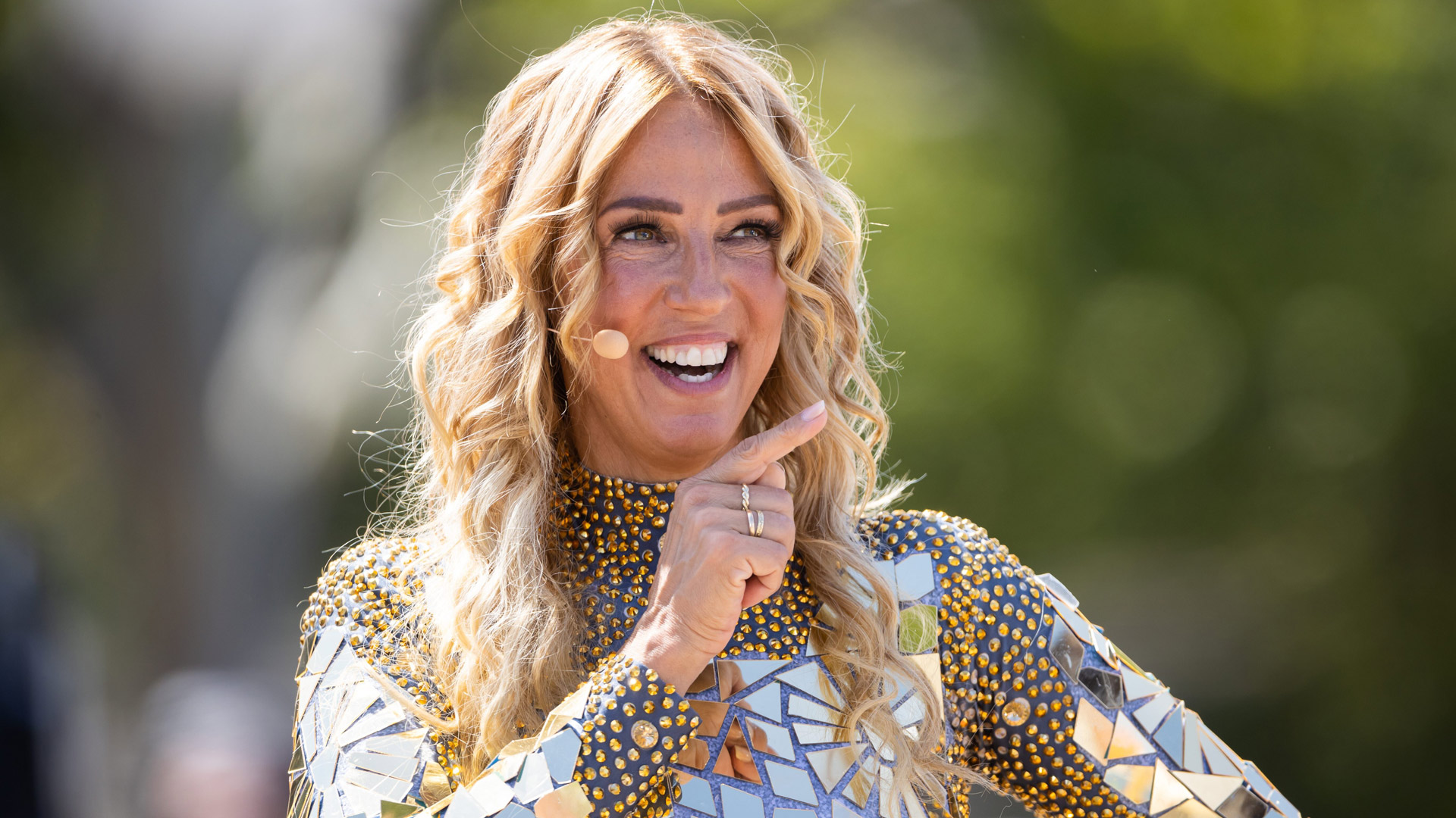 Auf dem Bild zu Kampf der RealityAllstars 2026 ist die Sängerin und Reality-TV-Teilnehmerin Loona fröhlich lächelnd zu sehen. Sie trägt ein auffälliges Glitzeroutfit mit goldenen und silbernen Spiegelelementen. Mit einem Headset-Mikrofon ausgestattet, zeigt sie mit dem Finger nach vorn. Die Stimmung ist gelöst und positiv, das Bild wurde im Freien aufgenommen.