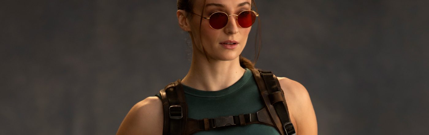 Auf dem Bild zur Tomb Raider Serie ist Sophie Turner als Lara Croft zu sehen. Sie trägt ein enges, dunkelgrünes Tanktop und ein taktisches Geschirr mit Schnallen. Ihre Haare sind zurückgebunden, einige Strähnen fallen locker ins Gesicht. Auffällig sind die runden, roten Sonnenbrillen, die ihrem entschlossenen Gesichtsausdruck eine coole Note verleihen.