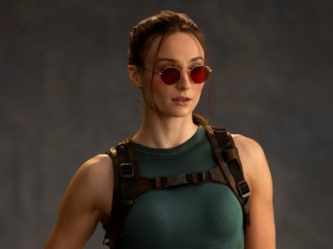 Auf dem Bild zur Tomb Raider Serie ist Sophie Turner als Lara Croft zu sehen. Sie trägt ein enges, dunkelgrünes Tanktop und ein taktisches Geschirr mit Schnallen. Ihre Haare sind zurückgebunden, einige Strähnen fallen locker ins Gesicht. Auffällig sind die runden, roten Sonnenbrillen, die ihrem entschlossenen Gesichtsausdruck eine coole Note verleihen.