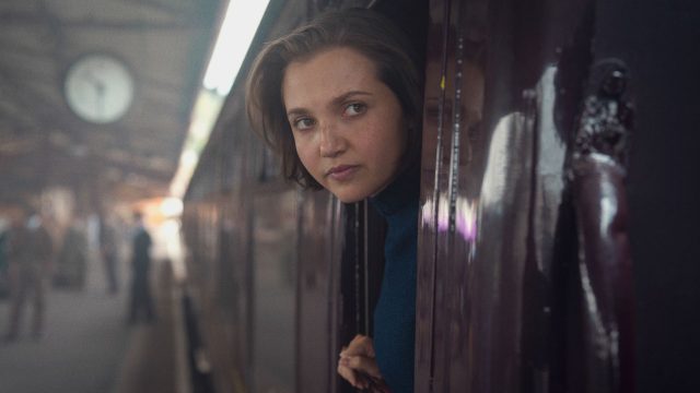 Mia McKenna-Bruce als Bundle schaut in einer Szene von "Agatha Christie's Seven Dials" aus einem Zugfenster heraus.