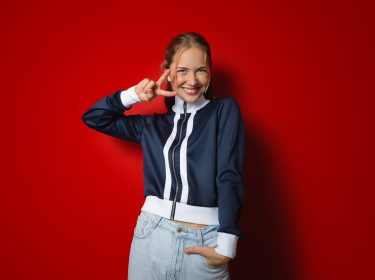 Zum Netflix-Angebot für Vodafone Mobilfunk-Kund:innen zeigt das Bild eine fröhliche junge Frau vor einem kräftig roten Hintergrund. Sie trägt eine sportliche dunkelblaue Jacke mit weißen Streifen und lächelt strahlend, während sie ein Victory-Zeichen mit der rechten Hand macht