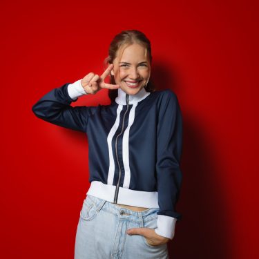 Zum Netflix-Angebot für Vodafone Mobilfunk-Kund:innen zeigt das Bild eine fröhliche junge Frau vor einem kräftig roten Hintergrund. Sie trägt eine sportliche dunkelblaue Jacke mit weißen Streifen und lächelt strahlend, während sie ein Victory-Zeichen mit der rechten Hand macht