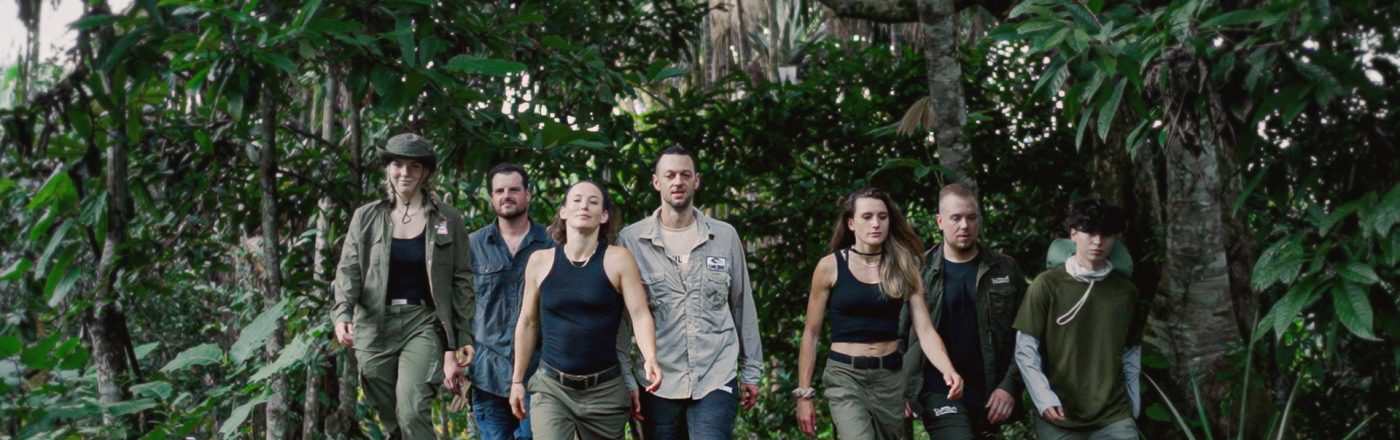 Auf dem Bild zu 7 vs. Wild Gewinner:innen sind sieben Teilnehmende im Dschungel zu sehen. Von links nach rechts: Jeannine Michaelsen, Joe Vogel, Fibii, Lucas „Avive“ Michels, Imke Salander, Kris Grippo und David Leichtle. Alle tragen Outdoor-Kleidung und wirken fokussiert und entschlossen, während sie durch dichtes Grün marschieren.