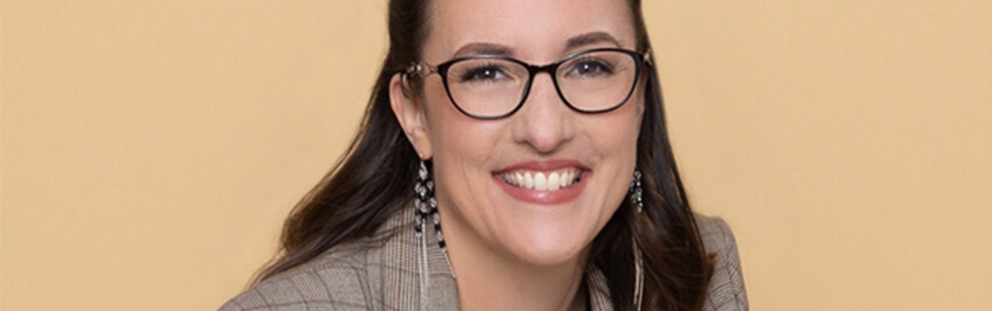 Auf dem Bild ist Abigail Owen, die Autorin der The Games Gods Play-Reihenfolge, vor einem beigefarbenen Hintergrund zu sehen. Sie trägt eine karierte Jacke, eine Brille und auffällige Ohrringe. Mit einem offenen Lächeln wirkt sie sympathisch und professionell – ein stimmiges Autorenportrait für ihre erfolgreiche Fantasyreihe.