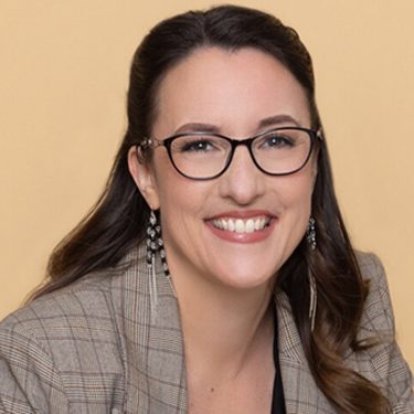 Auf dem Bild ist Abigail Owen, die Autorin der The Games Gods Play-Reihenfolge, vor einem beigefarbenen Hintergrund zu sehen. Sie trägt eine karierte Jacke, eine Brille und auffällige Ohrringe. Mit einem offenen Lächeln wirkt sie sympathisch und professionell – ein stimmiges Autorenportrait für ihre erfolgreiche Fantasyreihe.