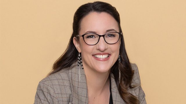 Auf dem Bild ist Abigail Owen, die Autorin der The Games Gods Play-Reihenfolge, vor einem beigefarbenen Hintergrund zu sehen. Sie trägt eine karierte Jacke, eine Brille und auffällige Ohrringe. Mit einem offenen Lächeln wirkt sie sympathisch und professionell – ein stimmiges Autorenportrait für ihre erfolgreiche Fantasyreihe.