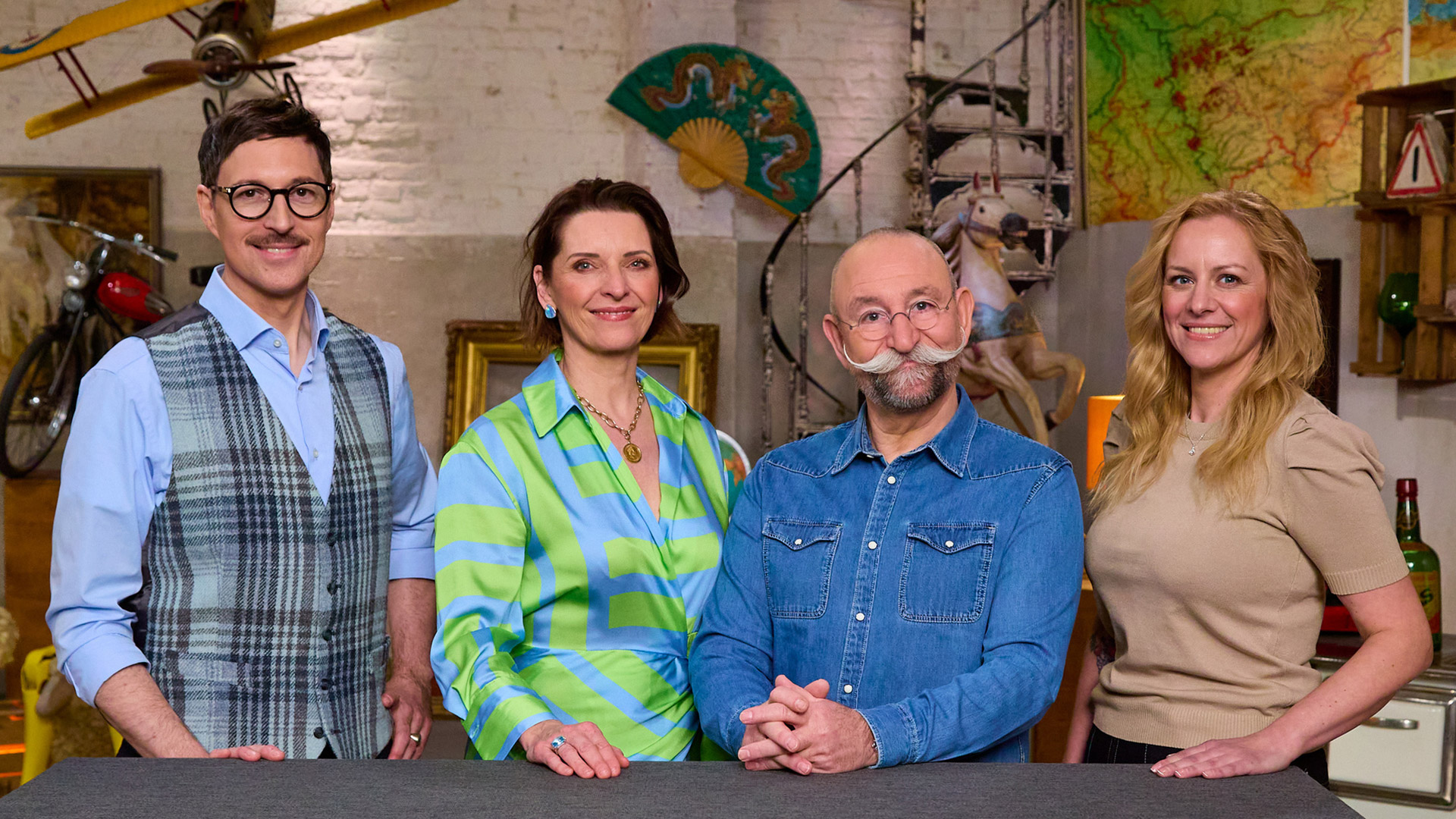 Auf dem Bild mit den Bares für Rares-Expert:innen stehen vier bekannte Gesichter der Trödelshow nebeneinander vor einem Studiotresen. Von links nach rechts sind Colmar Schulte-Goltz mit kariertem Anzug, Wendela Horz in einer blau-grünen Bluse, Moderator Horst Lichter mit rundem Brillenrahmen und Schnurrbart sowie Annika Raßbach in einem beigefarbenen Oberteil zu sehen. Die Atmosphäre wirkt offen und herzlich.