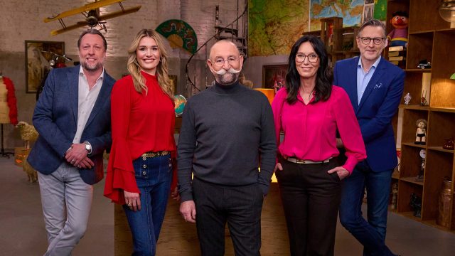 Auf dem Bild mit den Bares für Rares-Expert:innen stehen fünf bekannte Gesichter der Sendung lächelnd nebeneinander im Studio. Von links nach rechts sind Patrick Lessmann, Bianca Berding in roter Bluse, Moderator Horst Lichter mit markantem Schnurrbart, Heide Rezepa-Zabel in pinker Bluse und Detlev Kümmel im blauen Sakko zu sehen. Die Stimmung wirkt freundlich und professionell.