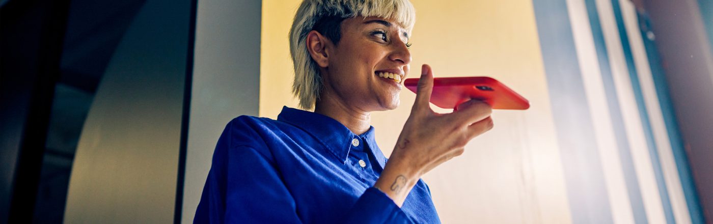 Eine lächelnde Person mit blondem Kurzhaarschnitt trägt ein leuchtend blaues Hemd und spricht in ein rotes Smartphone, als würde sie einen Sprachbefehl geben. Die Szene spielt sich in einem modernen Gebäude mit gelbem Hintergrund ab. Das Bild symbolisiert moderne Kommunikation und passt zum Thema Beste Sprachassistenten.