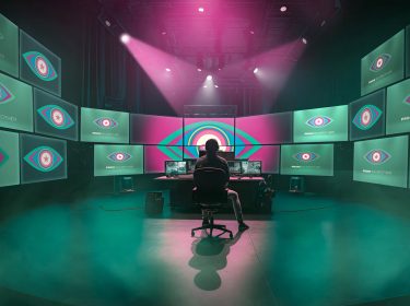 In diesem Bild zur Show Promi Big Brother sitzt eine Person in einem abgedunkelten Regieraum vor einer riesigen Monitorwand. Auf allen Bildschirmen ist das ikonische Auge-Logo von Promi Big Brother zu sehen. Die Szene ist in grünliches Licht und Nebel getaucht, darüber strahlen dramatische pinke Scheinwerfer – die Stimmung wirkt geheimnisvoll und überwachend.
