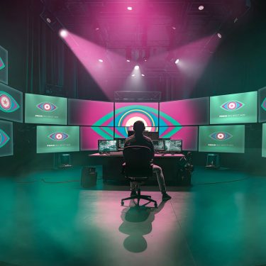 In diesem Bild zur Show Promi Big Brother sitzt eine Person in einem abgedunkelten Regieraum vor einer riesigen Monitorwand. Auf allen Bildschirmen ist das ikonische Auge-Logo von Promi Big Brother zu sehen. Die Szene ist in grünliches Licht und Nebel getaucht, darüber strahlen dramatische pinke Scheinwerfer – die Stimmung wirkt geheimnisvoll und überwachend.