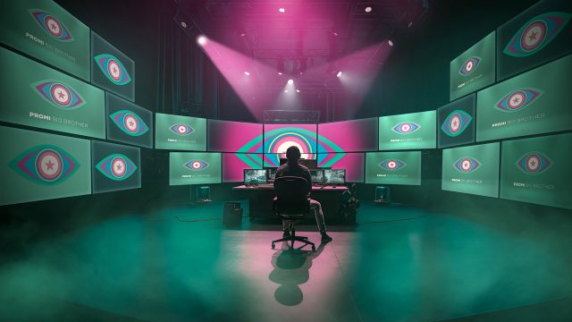In diesem Bild zur Show Promi Big Brother sitzt eine Person in einem abgedunkelten Regieraum vor einer riesigen Monitorwand. Auf allen Bildschirmen ist das ikonische Auge-Logo von Promi Big Brother zu sehen. Die Szene ist in grünliches Licht und Nebel getaucht, darüber strahlen dramatische pinke Scheinwerfer – die Stimmung wirkt geheimnisvoll und überwachend.