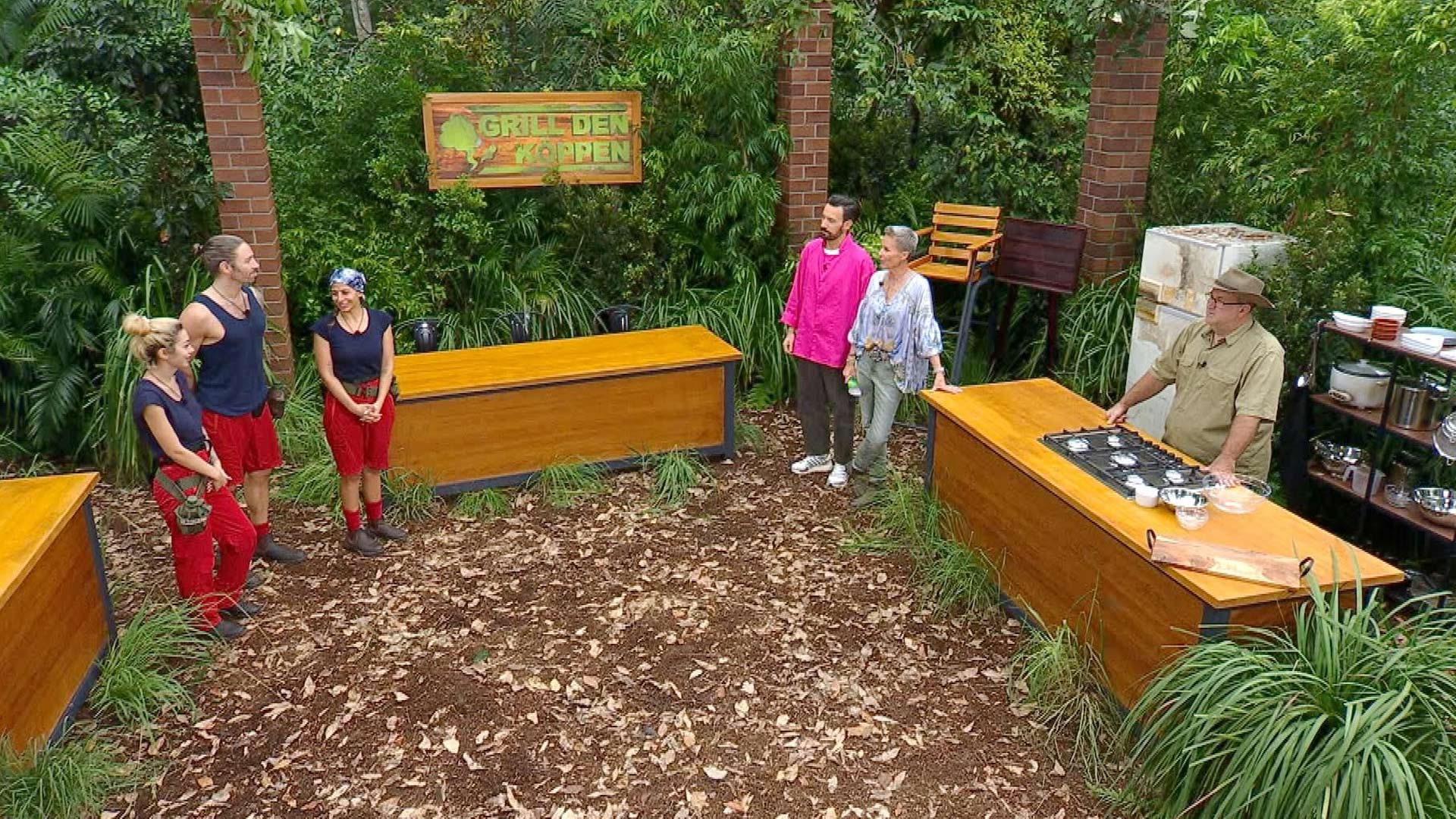 In dieser Szene aus dem Dschungelcamp 2026 stehen die Kandidat:innen Ariel, Gil Ofarim und Eva Benetatou links im Bild in roten Hosen und dunklen Shirts. Rechts stehen die Moderatoren Sonja Zietlow und Jan Köppen vor einem Holztresen, daneben ein weiterer Mann in Safari-Outfit. Im Hintergrund ist ein Schild mit der Aufschrift „Grill den Kopp(en)“ zu sehen, eingebettet in dichte Dschungelvegetation.